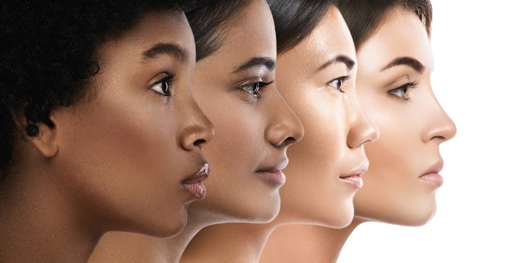 headshot of 4 women lined up looking away from the camera, arranged in order of light to dark skin tones with solid white background