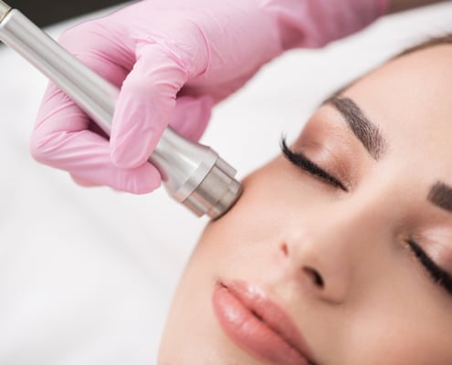 professional in pink gloves performing microdermabrasion on female patient’s face